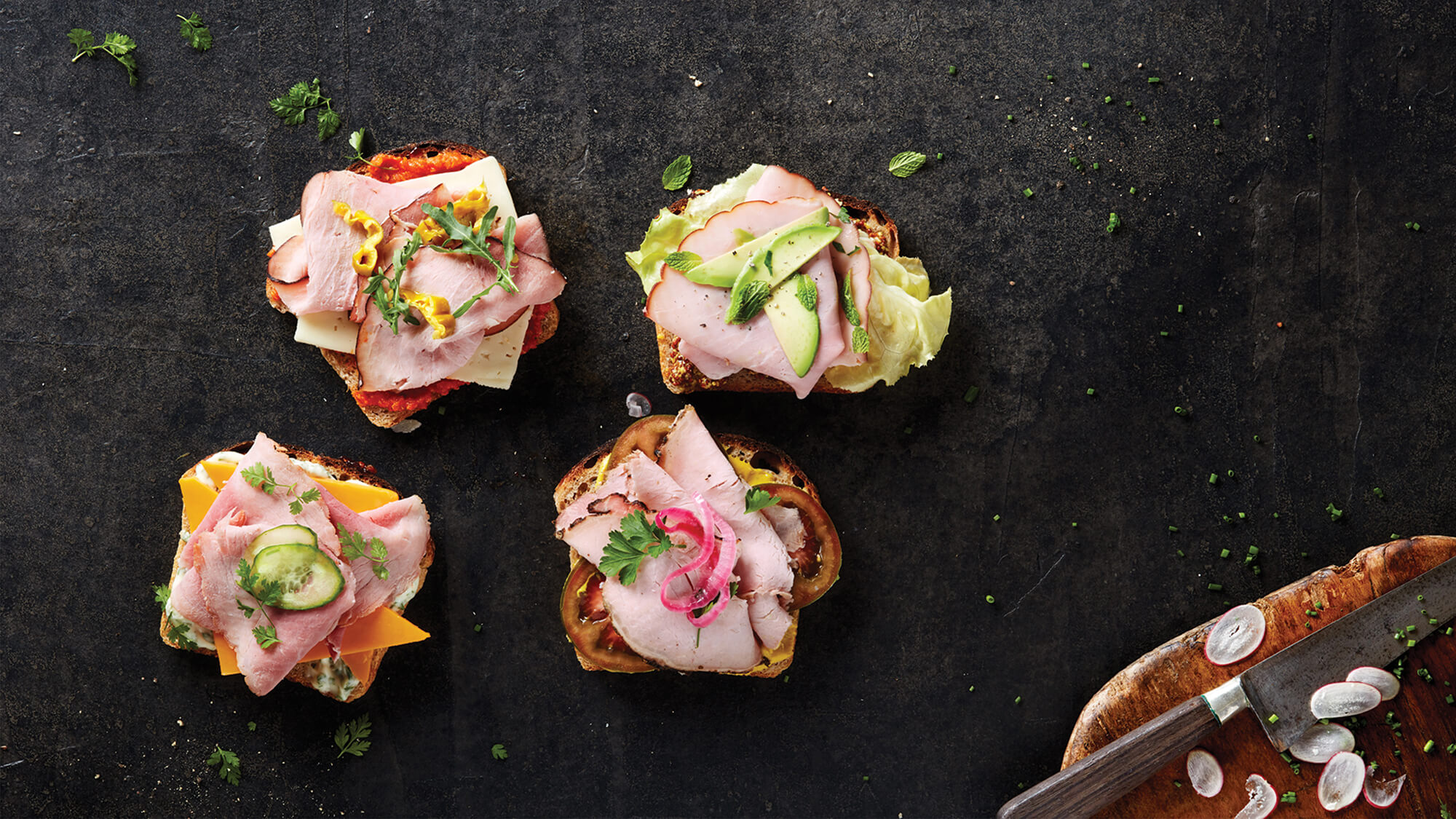 Group of four open-faced Columbus ham sandwiches with various toppings on a black slate