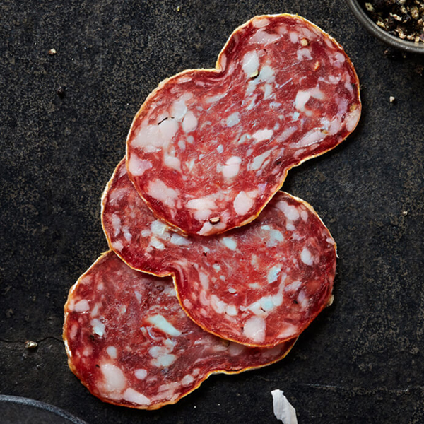 Flavorful Columbus Crespone Salami on black slate with a bowl of crushed peppercorn and a bowl of unpeeled garlic cloves