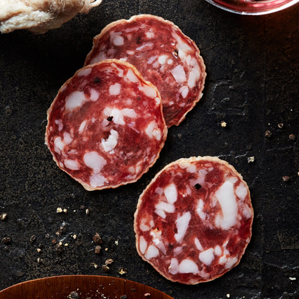 Sliced Columbus Felino Salami on black slate with a plate of crushed black peppercorns, dry aged Felino, and a glass of medium-bodied red wine