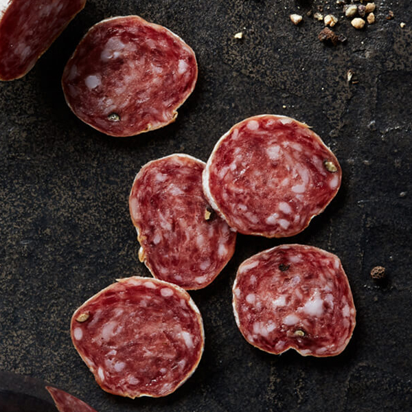 Fresh sliced Columbus Secchi Salami on black slate with crushed peppercorns and a glass of medium-bodied red wine